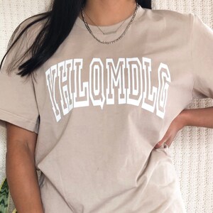 May include: A beige t-shirt with a white graphic print that reads "YHLQMDLG" in a bold, blocky font.