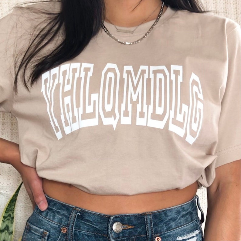 May include: A beige t-shirt with a white graphic that says "YHLQMDLG" in a large, bold font.
