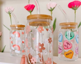May include: Three glass jars with bamboo lids and clear glass straws. The jars are decorated with pink and white flowers, hearts, and rainbow designs. One jar has candy hearts with Spanish text that reads "Bonita y Suelta" and "Bebecita".
