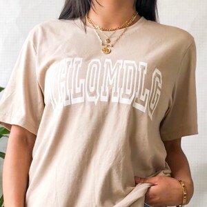 May include: A beige t-shirt with a white graphic that says "HLOMDLG" in a bold, blocky font.