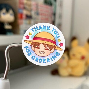 Puede incluir: Una pegatina redonda con un fondo acuarela azul, rosa y amarillo. La pegatina presenta una ilustración de dibujos animados de una persona con cabello castaño que lleva un sombrero de paja. El texto "Thank You For Ordering" está escrito en azul alrededor de la ilustración.
