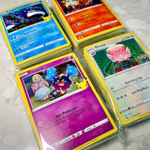 Puede incluir: Montones de cartas coleccionables de Pokémon, con personajes como Kyogre, Entei, Blissey y Cosmog. Las cartas tienen ilustraciones coloridas y texto, con un borde amarillo. Las cartas están empaquetadas en plástico transparente.