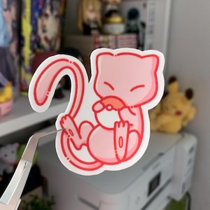 May include: A pink sticker featuring a cartoon-style Mew character. The sticker has a white outline and is held by tweezers. The character is in a curled position with its eyes closed. The sticker is on a white surface.