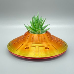 May include: A yellow and orange ceramic planter shaped like a flying saucer with a small green succulent plant growing inside.