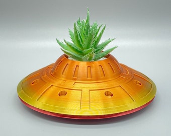 May include: A yellow and orange ceramic planter shaped like a flying saucer with a small green succulent plant growing inside.