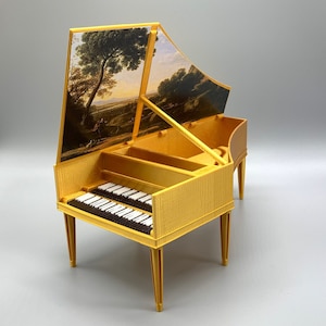 May include: A miniature gold-colored harpsichord with a landscape painting on the inside of the lid. The keyboard has black and white keys. The instrument has four slender legs and is set against a gray background.