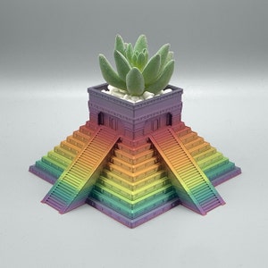 May include: A rainbow-colored ceramic planter shaped like a pyramid with a succulent plant growing inside. The planter has a small, rectangular building on top with a flat roof.