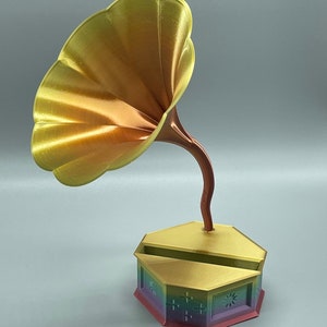 May include: A gold-colored, 3D printed, vintage-style gramophone with a rainbow-colored base. The horn is shaped like a flower and the base has a hexagonal shape with a decorative design.