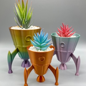 May include: Three rocket-shaped planters in different colors, each with a different succulent plant. The planters are made of a metallic material and have a gradient color effect. The succulents are green, blue, and red.