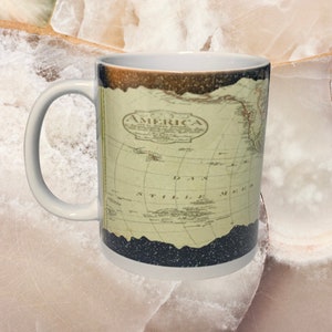 May include: White ceramic mug featuring a vintage map design with the word "AMERICA" at the top. The map is in shades of beige and brown, with black accents at the top and bottom of the mug.