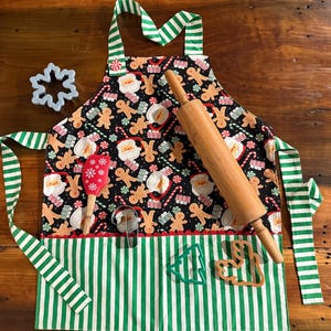 May include: A Christmas-themed apron with a black background featuring Santa Claus, gingerbread men, and candy canes. The apron has green and white striped trim and straps, along with a wooden rolling pin, a red spatula, and cookie cutters.