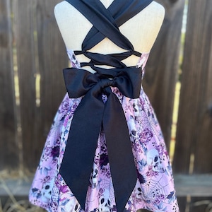 May include: A lilac-colored dress featuring a skull and floral print. The dress has a black bow and crisscross straps in the back. The skirt is flared, and the overall design is gothic-inspired.