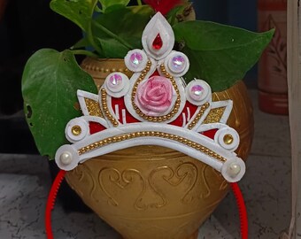 以下が含まれることがあります： ピンクの花、金色の縁取り、白いビーズが付いた白と赤のプリンセス冠ヘッドバンド。ヘッドバンドは赤で、上に羽根が付いています。