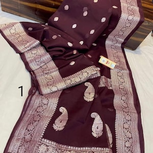 Puede incluir: Un sari de seda marrón con un borde plateado y un patrón de paisley. El sari tiene un brillo sutil y es perfecto para ocasiones especiales.