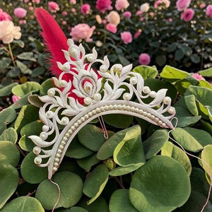 Op de afbeelding: Een witte tiara met parelaccenten en een rode veer. De tiara heeft een bloemenontwerp en is versierd met talrijke kleine parels. De achtergrond omvat groen gebladerte en roze rozen.