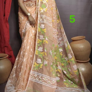 May include: A peach-colored saree with white and green floral patterns. The saree has a white border with a thin black stripe. The saree is draped over a person wearing a red blouse.