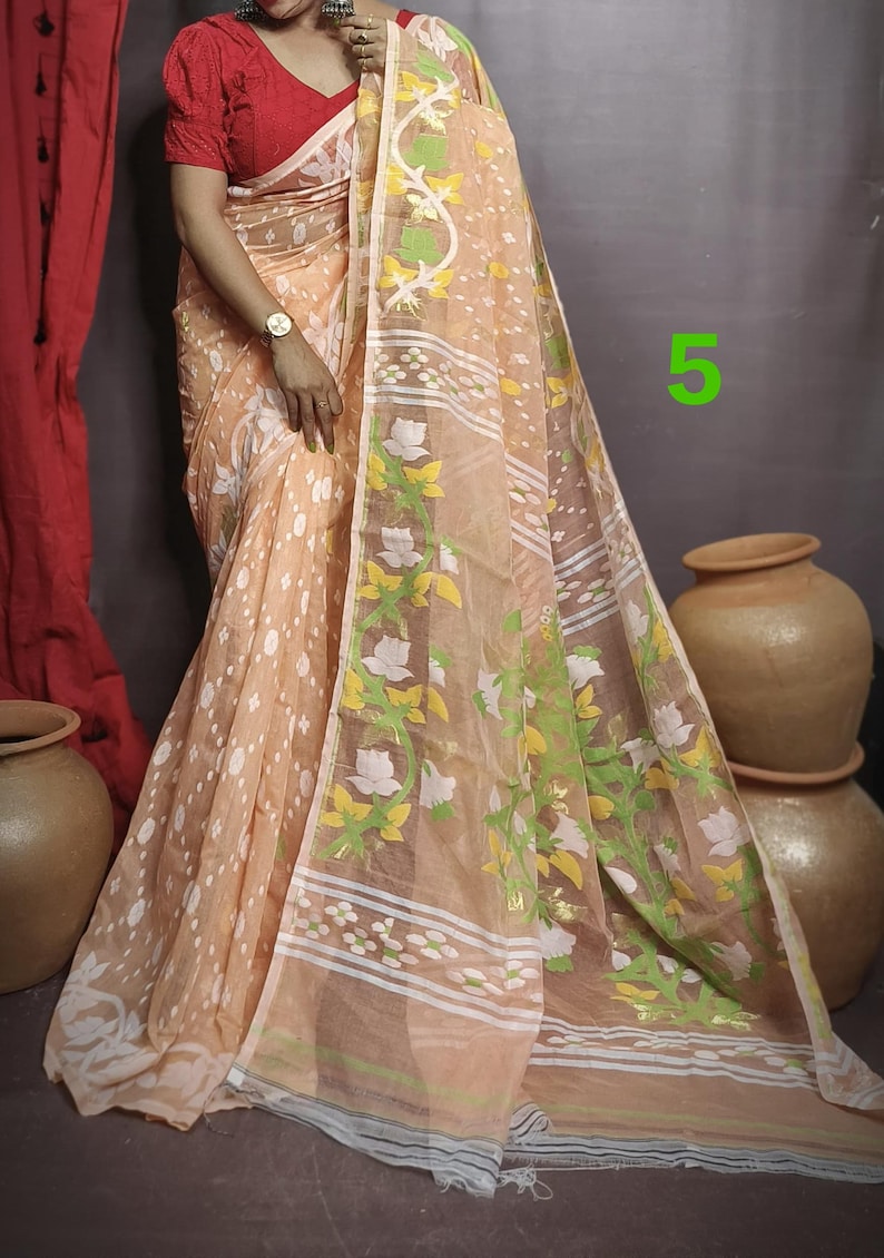 May include: A peach-colored saree with white and green floral patterns. The saree has a white border with a thin black stripe. The saree is draped over a person wearing a red blouse.