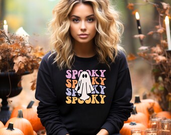 Może przedstawiać: Czarna bluza z nadrukiem białego ducha i tekstem "SPOOKY" powtarzanym w różnych kolorach. Bluzę nosi osoba z długimi, falującymi blond włosami. Osoba siedzi przed tłem jesiennych liści i dyni.