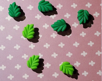 May include: A pink background with a white cross pattern. Seven green plastic leaves are arranged in a scattered pattern on the surface.