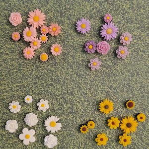May include: An assortment of colorful flower-shaped embellishments in various sizes. The flowers are in shades of pink, purple, yellow, and white, with yellow centers. The background is a green, grass-like texture.