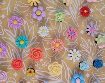 Op de afbeelding: Een verzameling kleurrijke bloemvormige bedels in verschillende maten en ontwerpen. De bedels zijn in tinten roze, geel, blauw, paars en rood. Sommige bedels hebben een smiley-gezicht. De achtergrond is een geel bloemenpatroon.