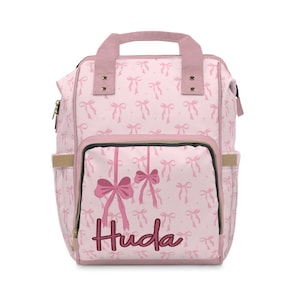 May include: Pink backpack with a repeating bow pattern. The front pocket features hanging bows and the name "Huda" in cursive. The bag has a top handle, two side pockets, and gold-colored zippers.