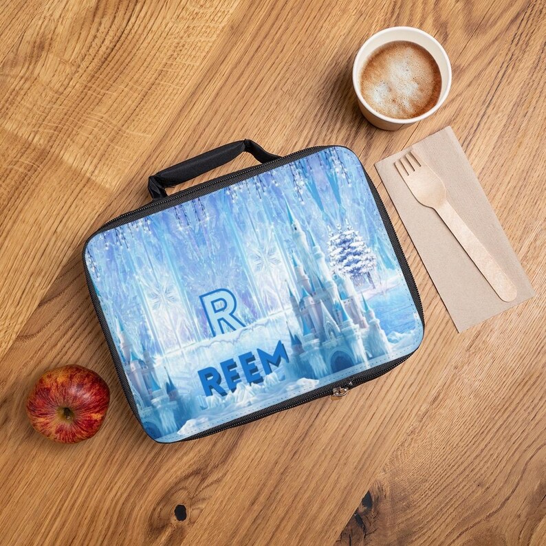 Puede incluir: Una bolsa de almuerzo azul y blanca con un dise&ntilde;o de castillo de invierno, con el nombre "REEM" en letras azules. La bolsa tiene un asa y cremallera negras. Una manzana, una taza de caf&eacute; y un tenedor de madera est&aacute;n sobre una superficie de madera.