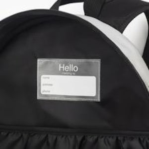 Puede incluir: Mochila negra con una franja plateada reflectante y un portaetiquetas transparente. El portaetiquetas tiene la palabra "Hello" impresa en la parte superior, con espacios para el nombre, la direcci&oacute;n y el n&uacute;mero de tel&eacute;fono.