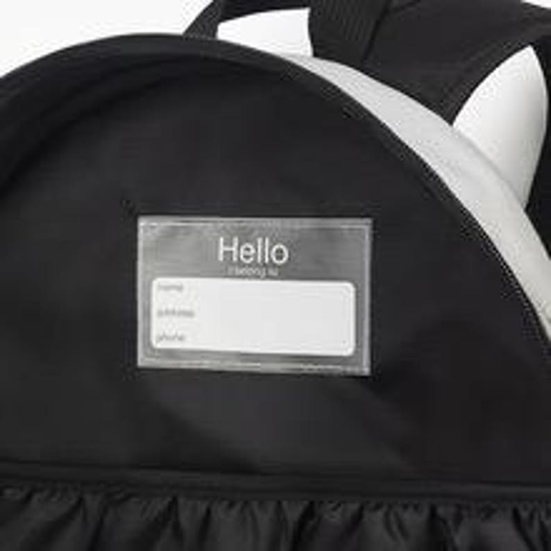 Puede incluir: Mochila negra con una franja plateada reflectante y un portaetiquetas transparente. El portaetiquetas tiene la palabra "Hello" impresa en la parte superior, con espacios para el nombre, la direcci&oacute;n y el n&uacute;mero de tel&eacute;fono.