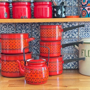 Puede incluir: Un conjunto de utensilios de cocina vintage de esmalte rojo con diseños florales naranjas, que incluye ollas, sartenes y una tetera. El conjunto se exhibe sobre una superficie de madera.