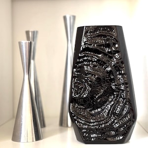 Può includere: Un vaso nero strutturato con un design organico unico è in evidenza, completato da tre candelabri argentati di diverse altezze. Il vaso ha una finitura lucida e i candelabri hanno un aspetto metallico liscio.