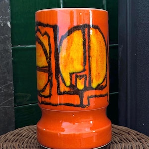 May include: A cylindrical orange ceramic vase with a flared base and a wide, rounded top. The vase features a bold, abstract design in yellow and black. The vase is sitting on a woven wicker surface.