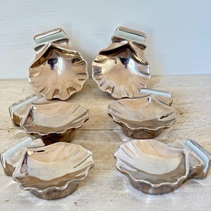Könnte beinhalten: Sechs silberfarbene, muschelförmige Schalen. Jede Schale hat einen gewellten Rand und einen kleinen, rechteckigen Akzent. Die Schalen sind auf einer hellen Holzoberfläche angeordnet. Die Schalen sind reflektierend und scheinen aus Metall zu sein.