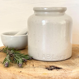 Può includere: Un vaso di ceramica grigio chiaro con corpo arrotondato e bordo leggermente svasato. Il vaso ha il testo "AB Lagny" in rilievo sulla base. Un piccolo mortaio e pestello bianchi sono sullo sfondo, insieme a un rametto di lavanda.