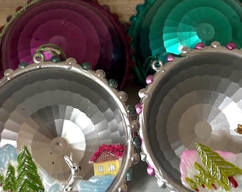 Op de afbeelding: Twee decoratieve kerstballen. Elke ornament heeft een zilverkleurige, facetgeslepen basis met een miniatuur winters tafereel binnenin. De scènes omvatten een huis, bomen en een konijn. De bovenkanten zijn paars en groen met kralenranden.