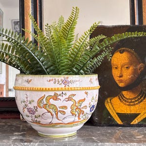 Könnte beinhalten: Weißer Keramiktopf mit handbemaltem Design von Vögeln und floralen Akzenten. Der Topf hat einen gelben Rand und ist mit grünen Farnpflanzen gefüllt. Der Topf steht auf einer Marmoroberfläche.