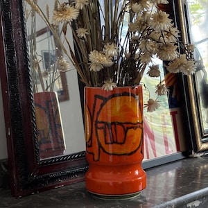 Può includere: Un vaso in ceramica arancione vibrante con un design astratto nero, contenente fiori secchi e spighe di grano. Il vaso è cilindrico con una parte superiore svasata e una base verde. Il vaso è alto circa 20 cm.