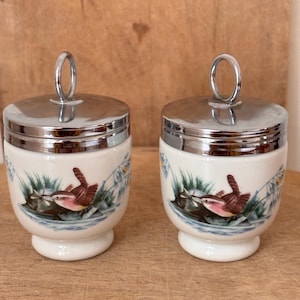 May include: Two white ceramic egg coddlers with silver-toned metal lids featuring a circular handle. Each coddler is decorated with a colorful illustration of a small brown bird perched among green reeds and blue water elements.