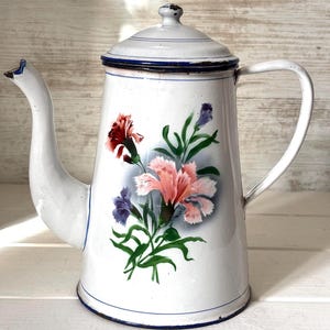 Könnte beinhalten: Weiße Emaille-Kaffeekanne mit floralem Design mit rosa, roten und violetten Blumen. Die Kanne hat einen blauen Rand und einen Deckel.