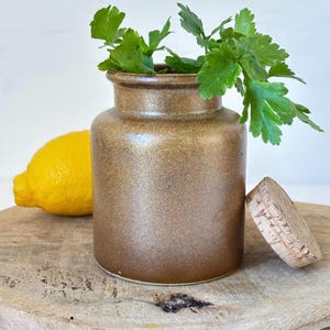 May include: A brown ceramic jar with a cork lid, filled with fresh green herbs. The jar is sitting on a wooden surface.