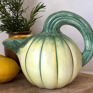 May include: A ceramic jug in the shape of a squash, featuring a green handle and spout. The jug is predominantly yellow with green vertical stripes. A sprig of rosemary in a brown pot and a lemon are in the background.