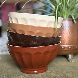 Op de afbeelding: Een stapel van vijf keramische kommen in verschillende bruintinten, van crème tot donkerbruin. De kommen hebben een geschulpte rand en zijn op elkaar gestapeld. De kommen staan op een groen tafelkleed.