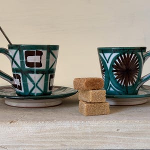 May include: Two ceramic coffee cups with matching saucers. The cups have green and white geometric patterns. One cup has a brown geometric pattern, the other has a brown floral pattern. There are three sugar cubes on a wooden surface.