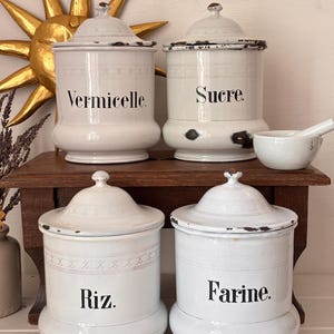 Może przedstawiać: Cztery białe ceramiczne pojemniki z pokrywkami, z napisami "Vermicelle", "Sucre", "Riz" i "Farine" czarnym tekstem. Pojemniki mają postarzany, vintage wygląd z dekoracyjnymi wykończeniami i są ustawione na drewnianej półce. Widoczny jest również mały moździerz i tłuczek.