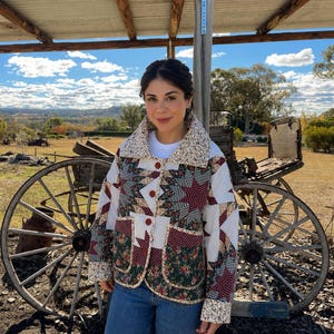 May include: A patchwork jacket with a floral collar and cuffs. The jacket features a mix of red, green, and white patterned fabrics, with a button-up front. The person is wearing blue jeans. The background includes a wooden wagon wheel and a rural landscape.