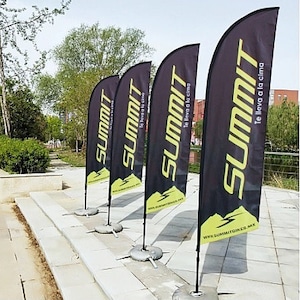 May include: Four black and yellow feather flags with the word "SUMMIT" and the text "Te lleva a la cima" in Spanish. The flags are displayed outdoors on a sidewalk.