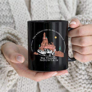 Op de afbeelding: Zwarte koffiemok met een wit ontwerp met een cartoon afbeelding van een achtbaanrit genaamd "Big Thunder Mountain". De tekst luidt "The wildest ride in the wilderness".