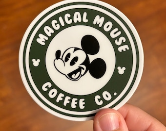 May include: A white round sticker with a green border. The sticker features a black and white cartoon image of Mickey Mouse with the text "Magical Mouse Coffee Co."