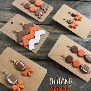 May include: Five pairs of handmade earrings featuring various designs inspired by the Cleveland Browns football team. The designs include footballs, helmets, and geometric shapes in brown and orange.