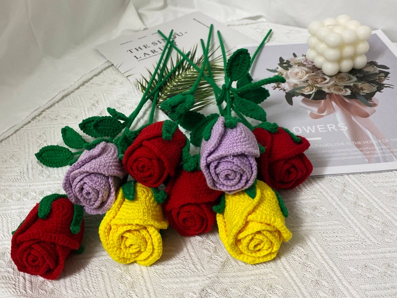 May include: A bouquet of crocheted roses in various colors, including red, yellow, purple, and pink. The roses are arranged in a vase and have green leaves.